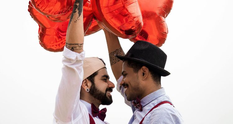 Two men embracing holding red heart shaped balloons.
